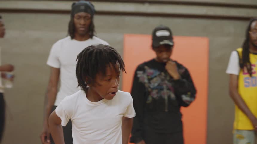 A talented young boy with dreadlocks dances with intense energy on an indoor basketball court as his friends and family watch