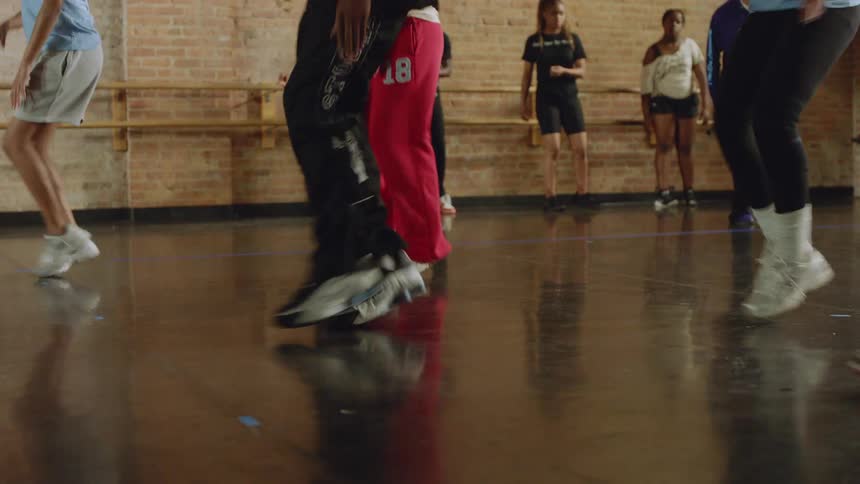 A group of dancers' feet move in unison as they practice a routine on a reflective studio floor in slow motion