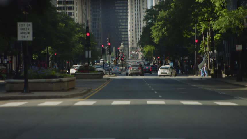 A pedestrian crosses a downtown city street with traffic moving between skyscrapers and trees