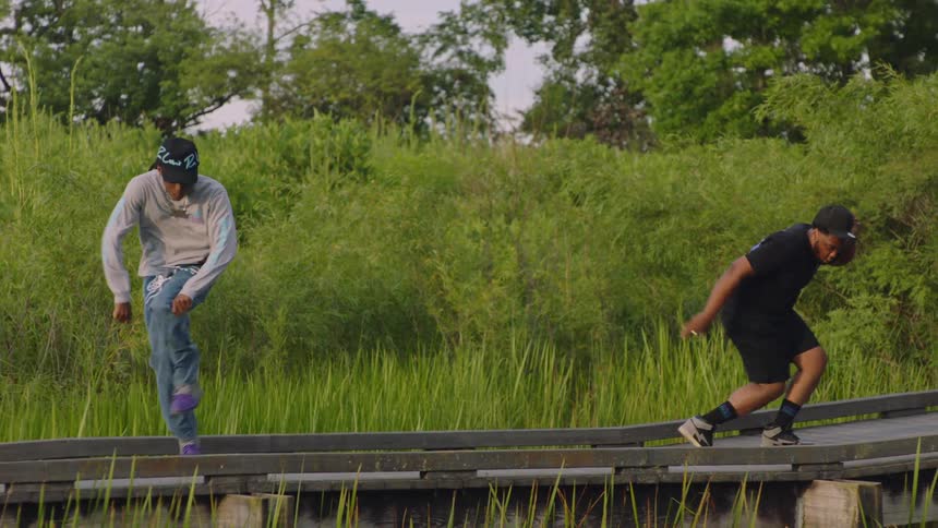 A group of diverse male dancers perform energetic hip-hop choreography in slow motion across a boardwalk in a lush green park