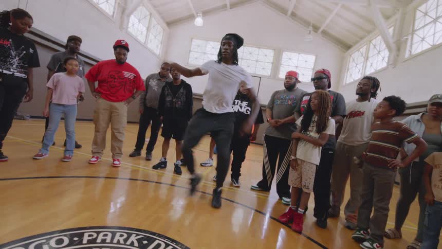 A talented footwork dancer showcases his impressive moves for a captivated crowd on a basketball court in a community center