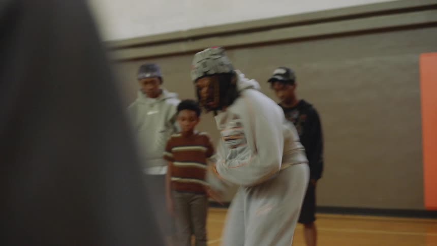 A young man performs energetic street dance footwork in slow motion for a group of onlookers in a gymnasium