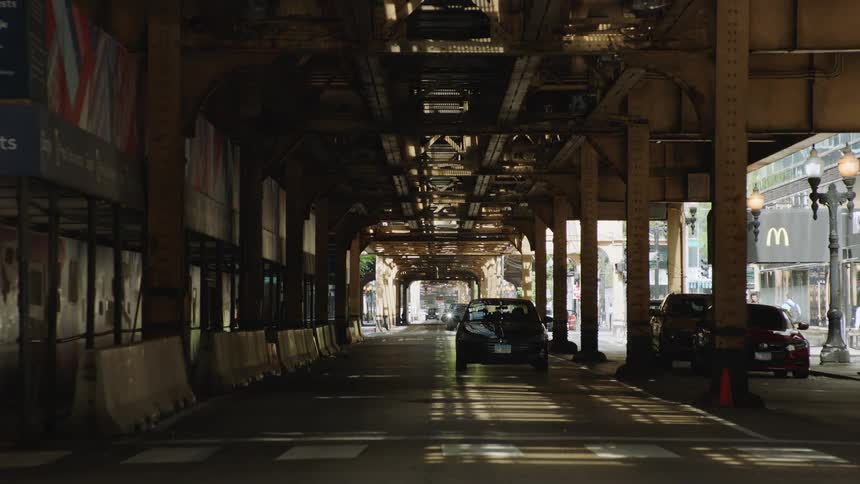 A black car slowly drives toward the camera on a quiet city street underneath the elevated train tracks