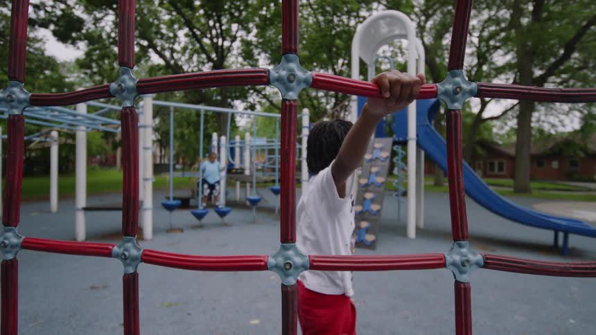 A young boy holds onto a red rope climbing net while playing at a park playground