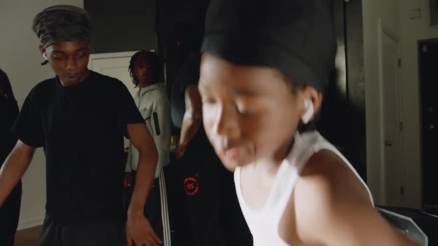 A young boy wearing a white tank top and earbuds energetically leads his friends in a hip-hop dance routine at home