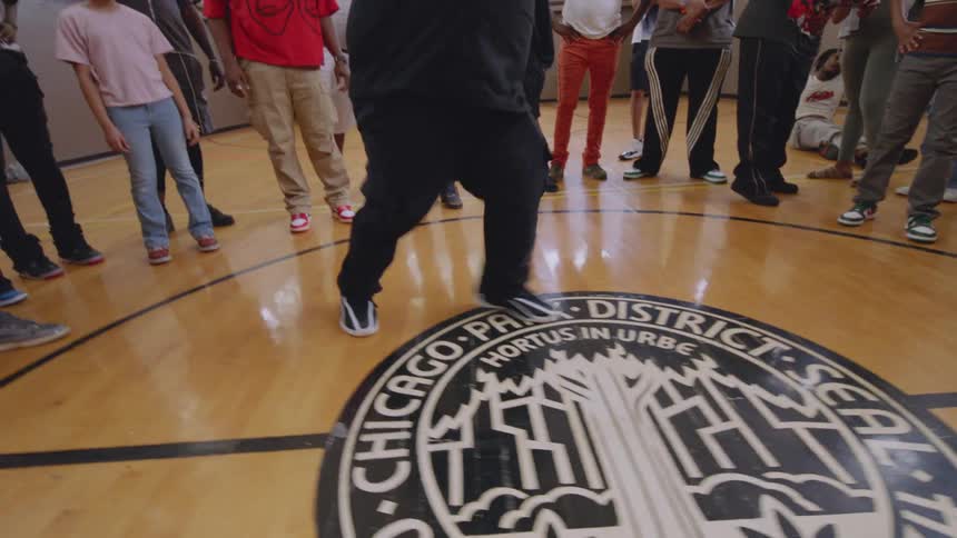 An energetic dancer with long braids performs intricate Chicago footwork in the center of a dance circle inside a gymnasium