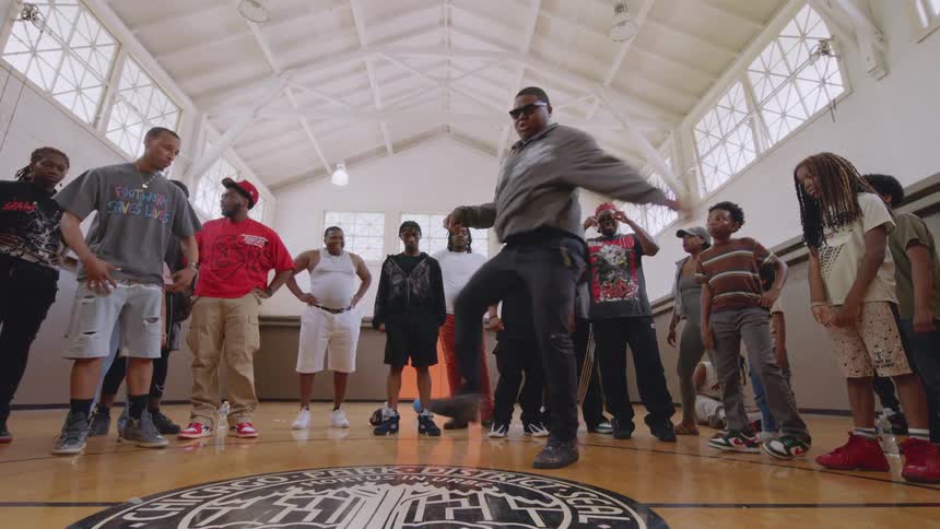 An energetic man performs an expressive street dance for a crowd of adults and children in a gymnasium dance circle