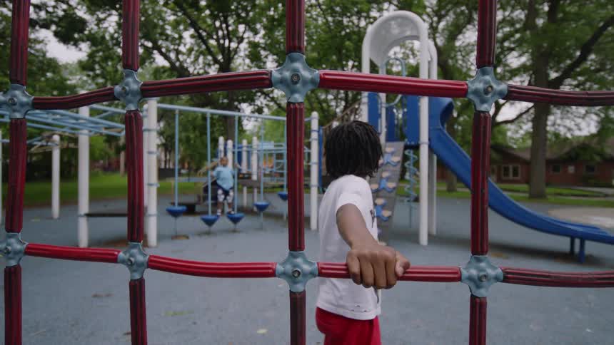A young boy with locs stands on a red rope climbing structure at a park playground, with another child playing in the background