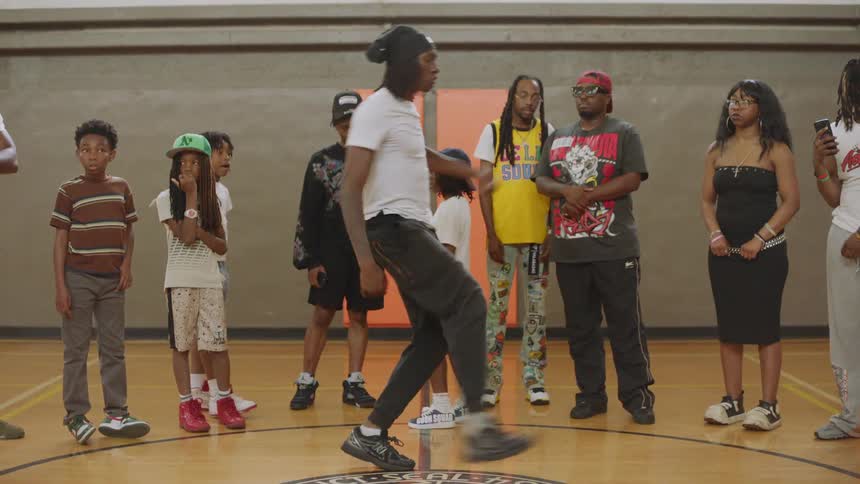 A young man with dreadlocks performs an energetic footwork dance for a crowd in a gymnasium