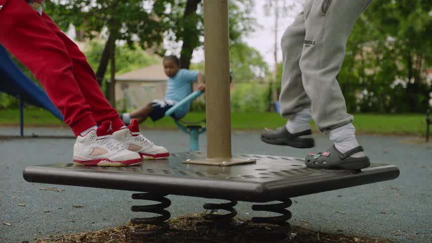 A slow-motion, low-angle shot shows the feet of two children bouncing on a spring rider at a local park