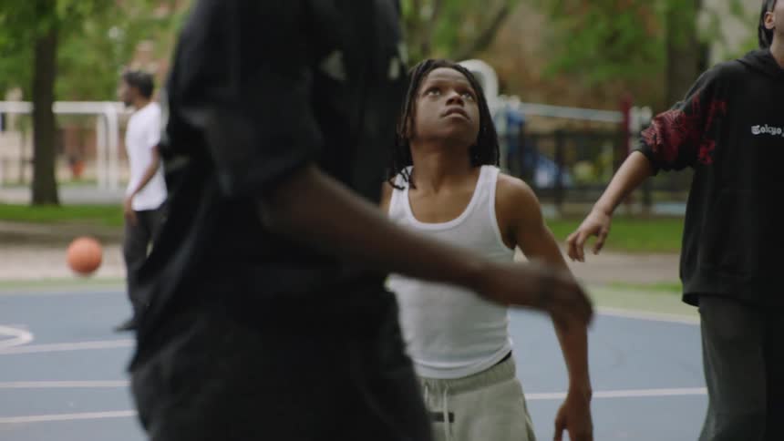 A young Black man with dreadlocks plays intense defense during a slow-motion pickup basketball game at a park