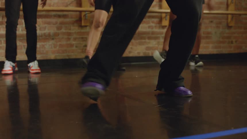 A tap dancer's feet in purple shoes move rhythmically during a dance class on a reflective studio floor