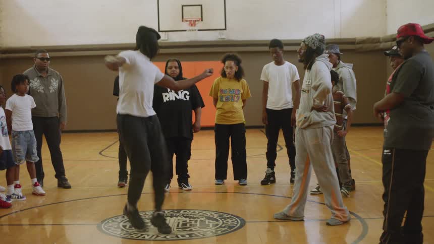 An energetic young man performs an intense krump dance routine in the center of a circle during a dance battle in a community gym