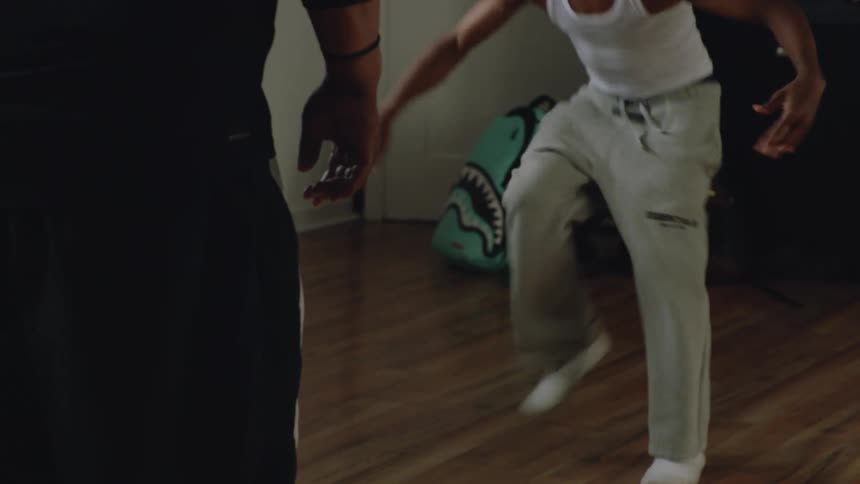 A talented young boy with dreadlocks and headphones performs an intense and energetic freestyle dance in a dimly lit room