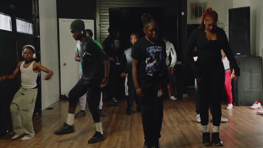 An energetic group of young Black dancers practices a hip-hop routine together in a dance studio