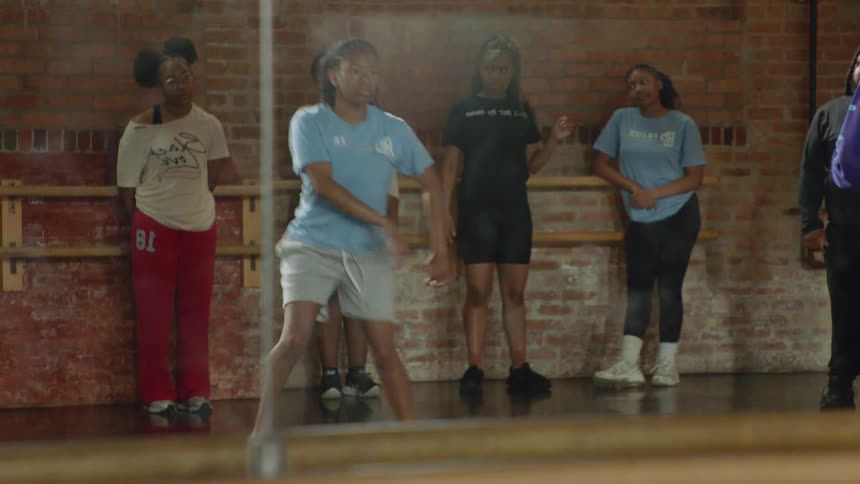 A young woman practices a hip-hop dance routine in front of a mirror as her friends watch in a studio