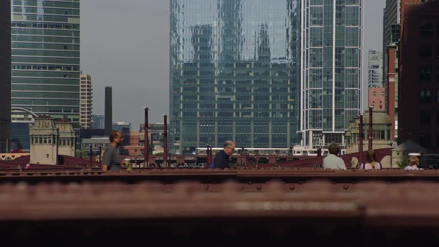 An elevated train travels across a bridge through downtown Chicago with towering glass skyscrapers and pedestrians in the background