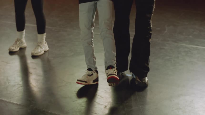 In a sunlit dance studio, a hip-hop instructor gently guides a young boy through a new dance move, with the camera tilting down to focus on their feet