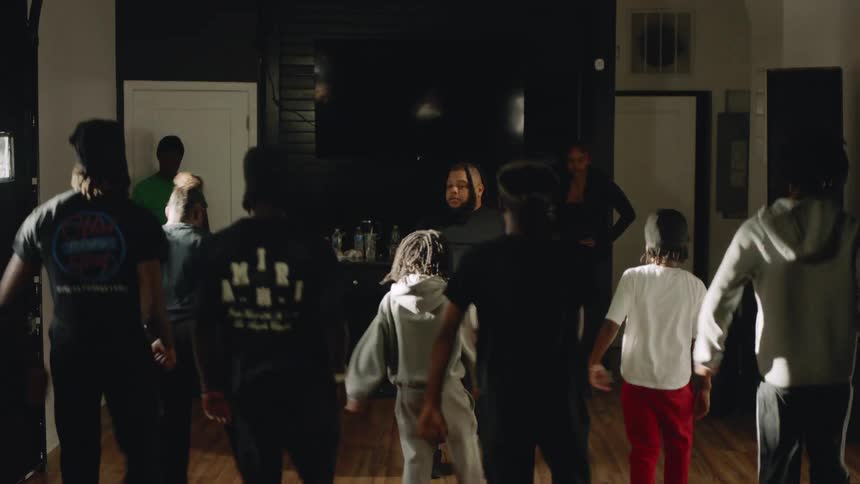 An instructor leads a diverse group of young people in a synchronized dance routine in a dimly lit studio