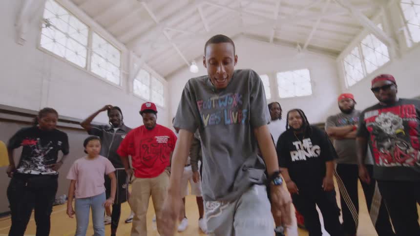 A man in a "Footwork Saves Lives" shirt performs a dynamic footwork dance routine in a circle of onlookers in a community center gym