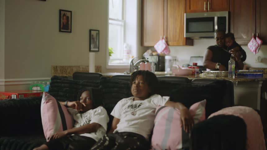 Two young boys relax on the couch watching television while their mother prepares food in the kitchen holding a baby