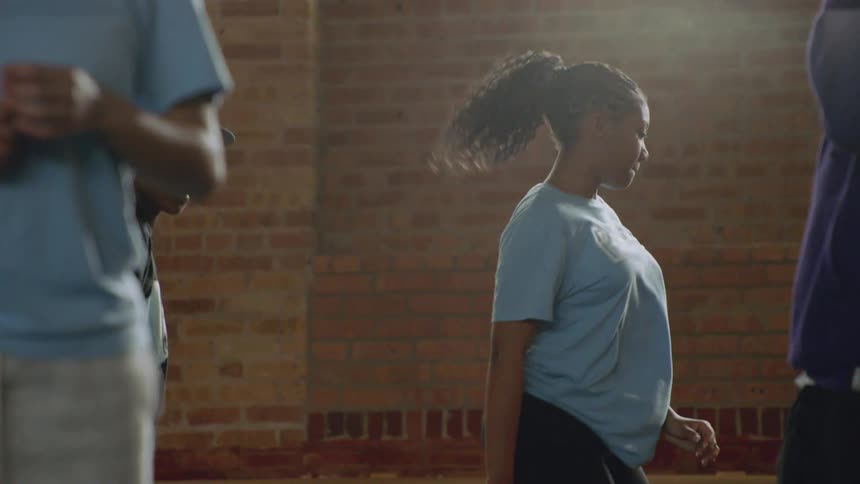 A young Black woman with a ponytail practices a dance routine in slow motion inside a sunlit studio with a brick wall