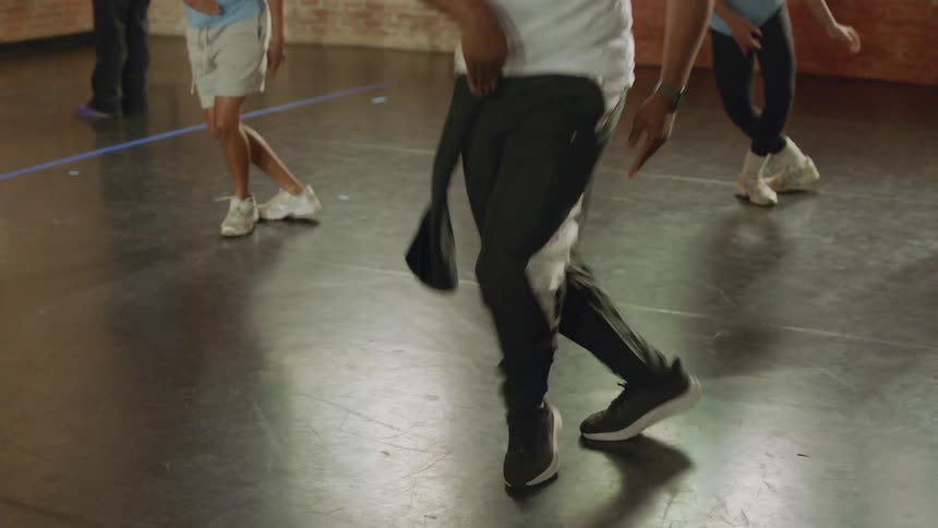 A male dance instructor leads a class of diverse students through a hip-hop routine in a spacious studio