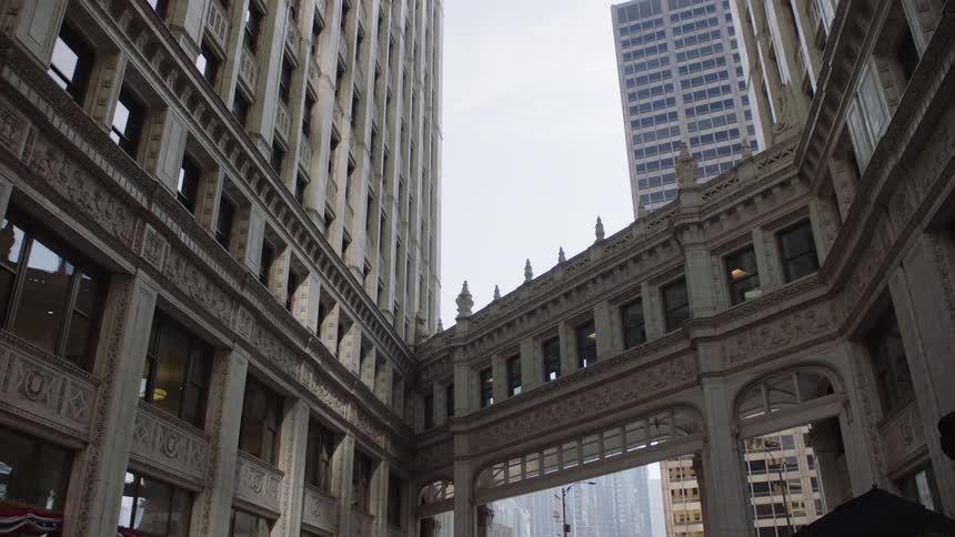 A static low angle view shows an ornate, historic skybridge connecting two buildings in a downtown city