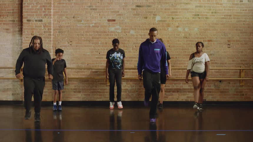 A dance instructor leads a diverse group of young students through a routine in a studio with a brick wall
