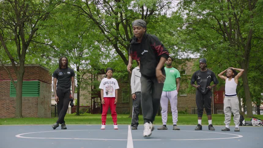 A young street dancer performs incredible, intricate footwork in slow motion for his friends on an outdoor basketball court