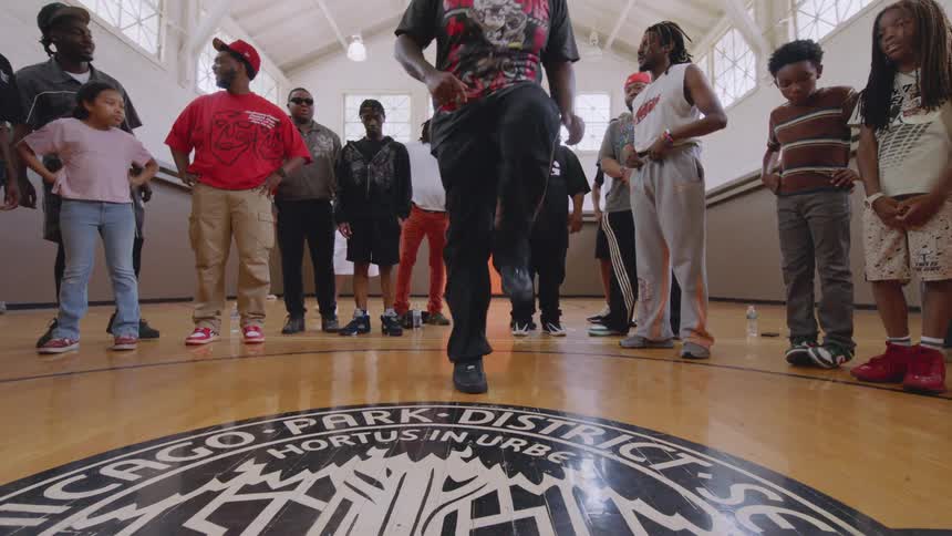 A skilled male dancer in sunglasses performs an energetic and intricate footwork routine for a crowd inside a Chicago community gymnasium