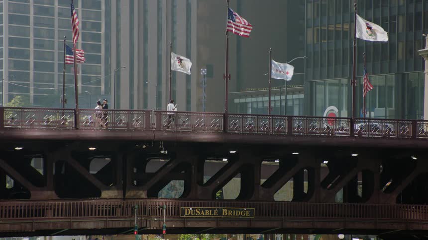 People walk across the historic DuSable Bridge in downtown Chicago as flags wave in the wind
