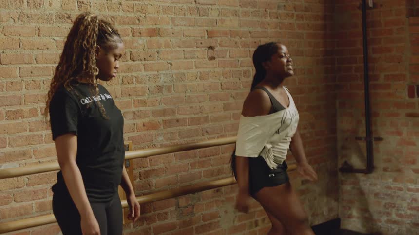 A young Black woman practices an energetic dance routine in slow motion while her instructor and classmates watch in a studio