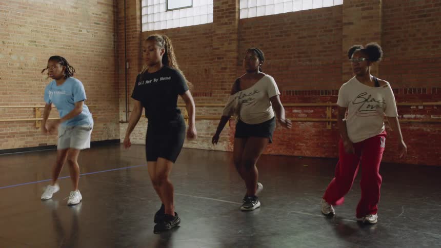 A group of four young Black women energetically practice a synchronized hip-hop dance routine in an urban studio