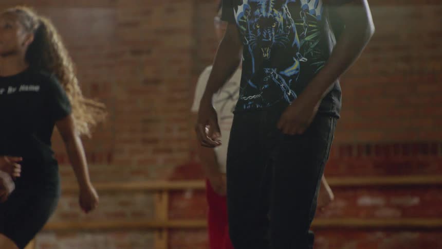 A group of dancers performs a synchronized routine in slow motion inside a studio with brick walls