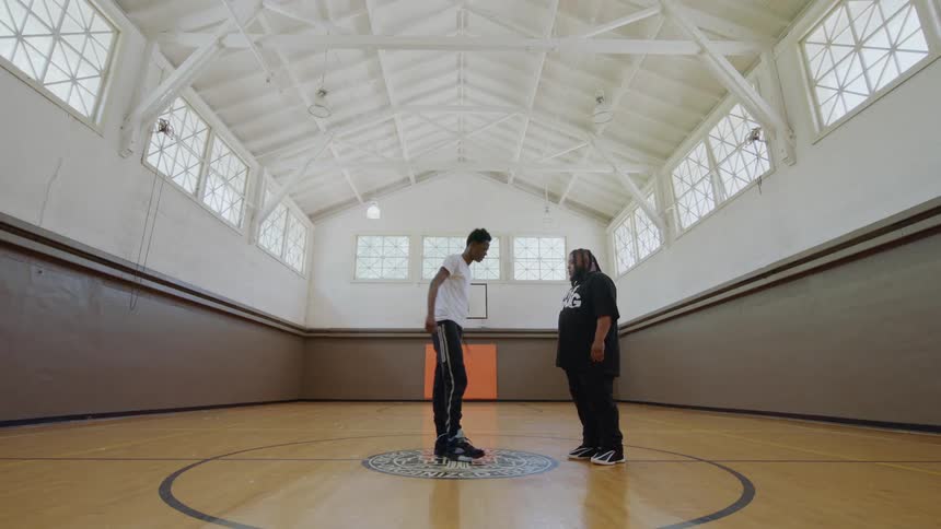A young man performs impressive street dance footwork for his mentor in the center of an empty gymnasium