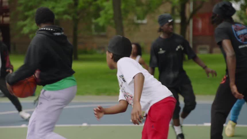 A group of young men play a competitive game of basketball in slow motion on an outdoor court in the park