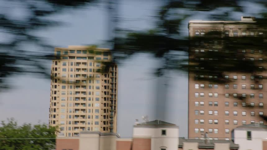 A fast-paced side-view tracking shot captures various city high-rise buildings through a blur of foreground trees and infrastructure