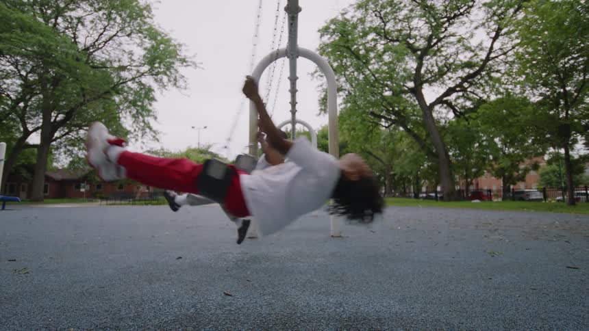 Two young friends swing joyfully on a playground in slow motion, their hair flying as they lean back and forth
