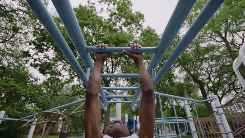 A low-angle, slow-motion shot captures a muscular young man doing leg raises on the monkey bars at an outdoor park