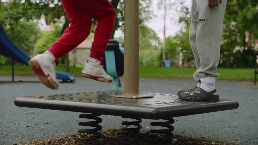 A low-angle view shows the legs and feet of two kids as they bounce on a spring-loaded platform at a playground