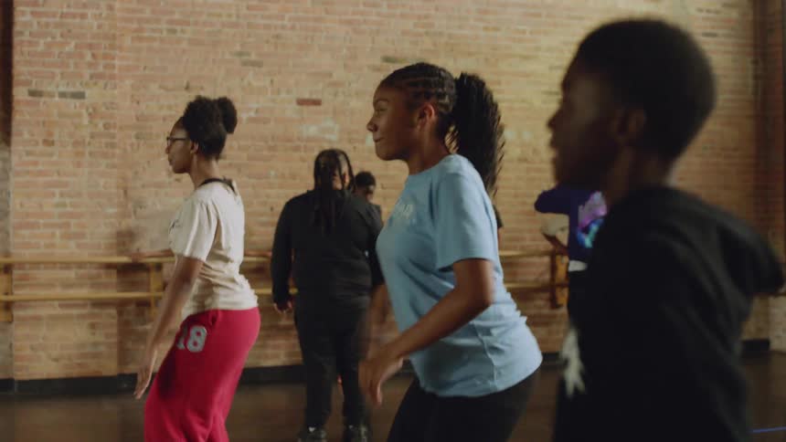 A diverse group of young dancers energetically practices a synchronized routine in slow motion in a sunlit studio with a brick wall