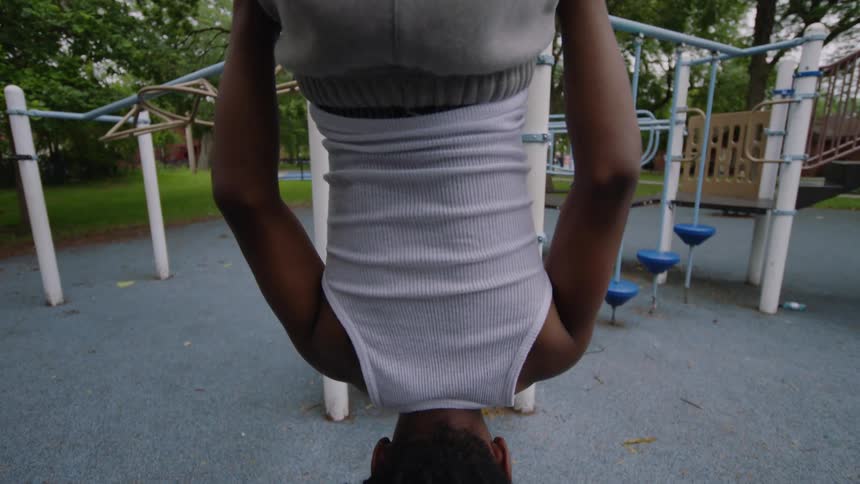 A slow tilt down reveals a young Black man with dreadlocks hanging upside down from the monkey bars at a playground
