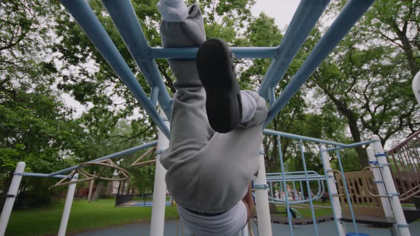 Slow motion low-angle shot of a young Black person with dreadlocks exercising and hanging upside down on monkey bars at a park