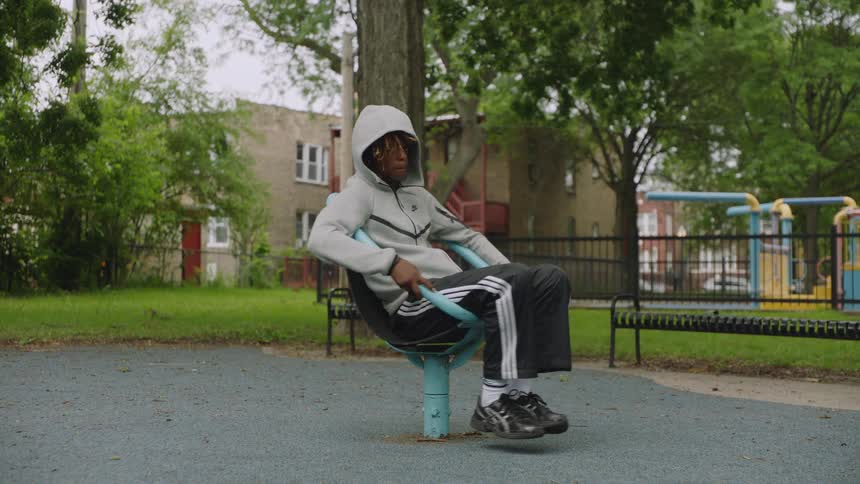 A happy young Black man in a hoodie spins around in slow motion on a playground merry-go-round