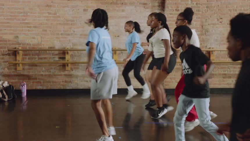 An instructor leads a diverse group of young students through a synchronized hip-hop routine in a dance studio in slow motion
