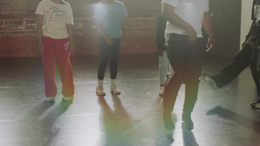 A dance instructor teaches a group of teens footwork in a sunlit studio, with the camera tilting down to focus on their movements