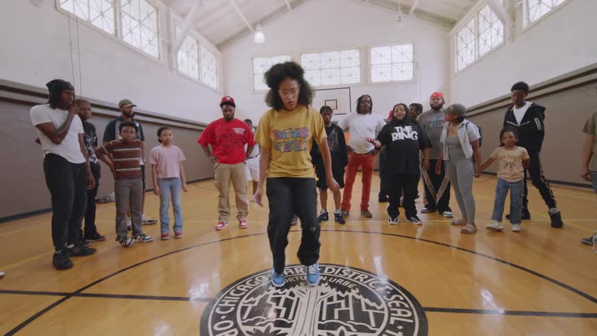 A dynamic dancer with an afro and glasses performs impressive Chicago footwork in the center of a gymnasium for an appreciative crowd
