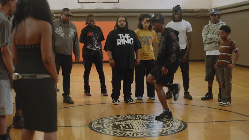A young street dancer performs impressive, fast-paced footwork for a crowd gathered on an indoor basketball court
