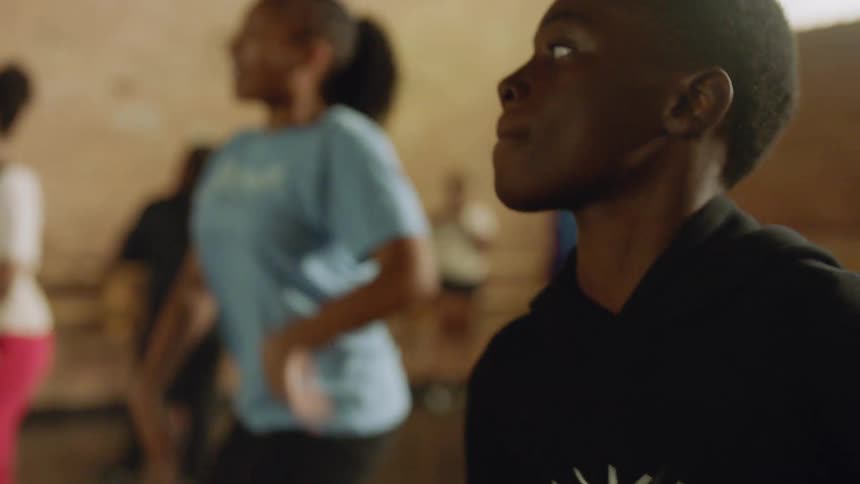A focused young Black person in profile, sweating and concentrating while participating in a dynamic group dance class